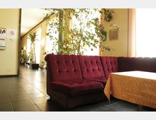 Ein Wartebereich mit einer gepolsterten roten Eckcouch, einem Tisch mit gelber Tischdecke und mehreren Zimmerpflanzen vor großen Fenstern, die natürliches Licht einlassen. Der Boden ist mit dunklen Fliesen bedeckt, und an der Wand hängt ein Rauchen-verboten-Schild.