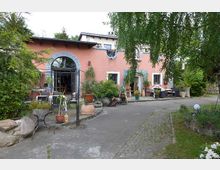 Ehemaliges Landhaus mit rosa Fassade und blauen Fensterläden, umgeben von Pflanzen und einem gepflasterten Hof. Im Vordergrund stehen Terrassenmöbel, Kübelpflanzen und Gestaltungselemente wie ein Brunnen.