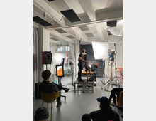Innenaufnahme eines hellen Fotostudios mit hohen Decken, sichtbaren Trägern und akustischen Paneelen, ausgestattet mit großen Softboxen, Scheinwerfern auf Stativen, Kameraaufbau und Kabeln. Im Hintergrund stehen ein Tisch vor einer dunklen Küchenzeile sowie große Fenster und Vorhänge.