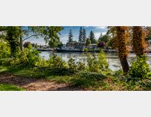 Ein Flussufer mit üppiger Vegetation im Vordergrund, darunter Bäume mit grünem und herbstlich verfärbtem Laub. Im Hintergrund sind mehrere Boote und ein Gebäude am gegenüberliegenden Ufer zu sehen, eingerahmt von hohen Nadelbäumen und weiteren Bäumen.