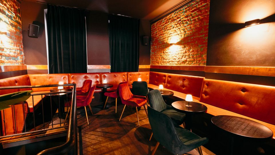 Gemütlicher Lounge- oder Barbereich mit langer, gepolsterter Leder-Sitzbank entlang der Wände, kleinen runden Tischen und gepolsterten Stühlen. Sichtbare Ziegelwände, dunkle Wandflächen mit Wandleuchten und schwere Vorhänge prägen die gedämpfte Innenraum-Atmosphäre.