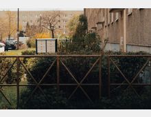 Wohngebiet mit einem Reihenhaus auf der rechten Seite, umgeben von Hecken und Pflanzen. Im Vordergrund ein Holzzaun, dahinter Briefkästen und dahinter parkende Autos sowie ein Wohnblock.