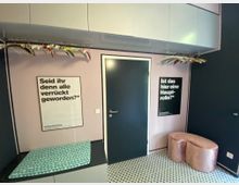 Ein kleiner, modern gestalteter Raum mit rosa und dunkelblauen Wänden. Es gibt eine dunkelblaue Tür in der Mitte, flankiert von zwei Plakaten mit schwarzem Text auf weißem Hintergrund, einem offenen Kleiderbügelhalter mit bunten Bügeln, sowie einer gepolsterten, rosafarbenen Sitzbank und einer schwarz-weißen Fliesenbodengestaltung.