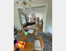 Großzügiger Wohnraum in einem Altbau mit Parkettboden und weißen Wänden. Im Vordergrund befindet sich ein Esstisch mit Holzstühlen und einem Blumenstrauß, während der Raum im Hintergrund ein Wohnzimmer mit Sofas, Regalen und einem großen bunten Kronleuchter zeigt.