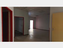 Leerer Innenraum mit einem hellen Laminatboden, weißen Wänden und einer Deckenverkleidung aus Rasterelementen. Die Türrahmen sind rot, eine gelbe Tür steht offen, und durch eine geöffnete Tür im Hintergrund fällt Tageslicht in den Raum.