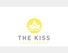 Logo mit gelbem Kreis, darin ein stilisiertes Segelboot über Wellen. Darunter in grauer und gelber Schrift die Worte „THE KISS“ und „KLOSTERSEE“ mit wellenförmigen Linien.