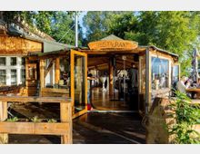 Holzrestaurant mit offenem Eingang und überdachtem Innenbereich, umgeben von einer Terrasse mit Holzmöbeln. Im Hintergrund befinden sich Bäume und Sonnenschirme, die eine Außenbewirtung andeuten.