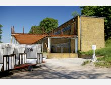 Ein einfaches, rechteckiges Gebäude aus gelblichen Ziegelsteinen mit Rostspuren an einer Metallüberdachung, umgeben von Grünflächen. Im Vordergrund stehen auf einem Anhänger mehrere weiße Strandkörbe mit Nummern; rechts befindet sich ein Schild mit der Aufschrift "Bootsverleih".