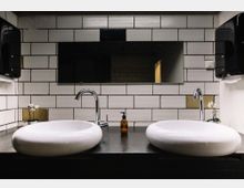 Moderne Badezimmeransicht mit zwei weißen Waschbecken auf einer dunklen Ablage, verchromten Armaturen und einer Rückwand aus weißen rechteckigen Fliesen. Im Hintergrund befindet sich ein breiter Spiegel, und auf beiden Seiten der Waschbecken stehen kleine Dekorationen sowie ein Seifenspender.