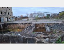 Verlassenes Industriegelände mit einer alten, teils eingestürzten Backsteinstruktur und einem offenen, mit Wasser gefüllten Graben; im Hintergrund stehen ein leerstehendes Gebäude und weitere Lagerhallen mit Bäumen und bewachsenem Boden.