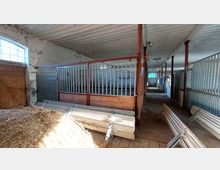 Innenansicht eines Pferdestalls mit Boxen aus Holz und Metallgittern, roten Metallstützen und einem weißen Holzdach. Im Vordergrund liegen Holz- und Metallteile sowie ein Strohhaufen, rechts und links sind Stalltrennwände sichtbar.