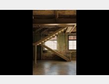 Industrie-innenraum mit einer modernen Metalltreppe, die diagonal verläuft. Die Wände sind aus unverputztem Ziegelmauerwerk, teils grün gestrichen, mit zwei großen Fenstern, die Tageslicht hereinlassen.