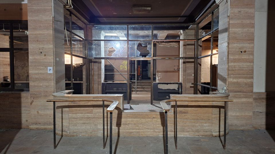 Verlassener Innenraum einer ehemaligen Empfangs- oder Kassenhalle mit zwei schmalen Tresen im Vordergrund und einer verglasten Trennwand dahinter. Wände und Säulen sind mit hellbraunem Stein verkleidet, einige Fensterscheiben sind beschädigt, und im Hintergrund führen Stufen in einen weiteren Bereich.