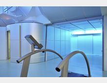 Ein moderner Raum mit blauer Beleuchtung, einer abgerundeten weißen Wand und einer Glastür. Im Vordergrund befindet sich ein Laufband mit integriertem Bildschirm.