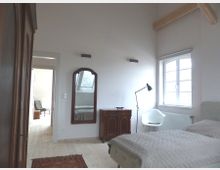 Schlafzimmer mit hellem Holzfußboden, einer Wand mit einem großen Fenster und einem Dachbalken. Ein dunkler Holzschrank, ein Wandspiegel, eine Kommode sowie ein Bett und ein moderner Stuhl prägen den Raum.