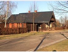 Holzhaus mit Satteldach, umgeben von Bäumen und niedrigen Hecken, an einem gepflasterten Weg in einer parkähnlichen Umgebung. Im Hintergrund sind kahle Bäume und ein klarer Himmel zu sehen.