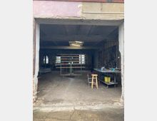 Geöffneter Eingangsbereich einer alten Werkstatt mit bröckelnder Fassade. Im Inneren sind ein Arbeitstisch, ein Hocker, ein Holzrahmen auf Rollen und verschiedene Werkzeuge sichtbar; die Wände sind teilweise unverputzt.