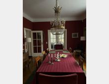 Esszimmer mit burgunderroten Wänden und einem langen Esstisch, der mit einer roten Tischdecke, Kerzenhaltern und einer Obstschale dekoriert ist. Ein prunkvoller Kristallleuchter hängt von der Decke, und im Hintergrund sind weiße Doppelflügeltüren zu sehen, die zu einer Terrasse führen.