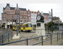 Straßenbahnhaltestelle in Alt-Köpenick mit gelber Straßenbahn, einer historischen Standuhr auf einem Sockel und mehrstöckigen Gebäuden mit Klinkerfassaden im Hintergrund. Im Vordergrund sind Fahrradbügel sowie ein gepflasterter Bürgersteig zu sehen.