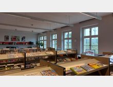 Innenraum einer Bibliothek oder Mediathek mit langen Regalen, die Zeitschriften, DVDs und Bücher enthalten. Große Fenster lassen Tageslicht herein, und an der Wand sind Regale mit Büchern sowie Bilder mit Naturmotiven zu sehen.