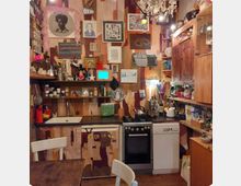 Kleine, kunstvoll dekorierte Küche mit bunten Holzpaneelen an den Wänden. Regale, Schränke und der Kühlschrank sind mit Gemälden, Postkarten und kleinen Gegenständen geschmückt; im Raum befinden sich ein Gasherd, eine Spüle, und ein Tisch mit weißen Stühlen.