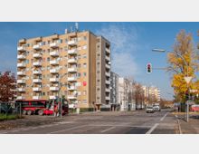 Hochhaus in einer städtischen Wohngegend mit gelblicher Fassade und mehreren Balkonen, umgeben von einer Straßenecke mit Ampeln und herbstlich gefärbten Bäumen. Im Vordergrund sind ein LKW sowie ein Gehweg sichtbar.