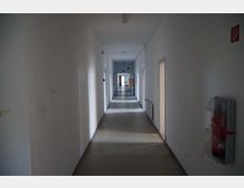 Ein langer, schlichter Korridor mit weißen Wänden und grauem Boden, beleuchtet durch indirektes Tageslicht. An der rechten Wand ist ein Feuerlöscher angebracht, und mehrere Türen führen zu angrenzenden Räumen.