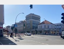 Straßenecke in einer Stadt mit mehreren Gebäuden im Hintergrund, darunter ein mehrstöckiges Bürogebäude mit großen Glasfenstern und ein ebenerdiges Eckgebäude mit roten Dachziegeln, das ein Fielmann-Geschäft beherbergt. Im Vordergrund eine belebte Kreuzung mit Autos, Fußgängern und Ampeln.