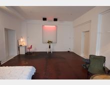 Ein schlicht eingerichteter Raum mit weißen Wänden und dunklem Holzboden. Im Vordergrund befindet sich ein Bett mit weißer Decke, ein Campingstuhl und ein kleiner Tisch; an der hinteren Wand steht ein weißer Beistelltisch mit einer Vase und Blumen unter einem leuchtenden Kunstobjekt mit rotem Licht.