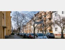 Straßenszene in einem Wohngebiet mit mehrstöckigen, hellen Gebäuden auf beiden Seiten und kahlen Bäumen entlang des Gehweges. Im Vordergrund steht ein Straßenschild mit der Aufschrift „Breitkopfstraße“, während Autos am Straßenrand parken.