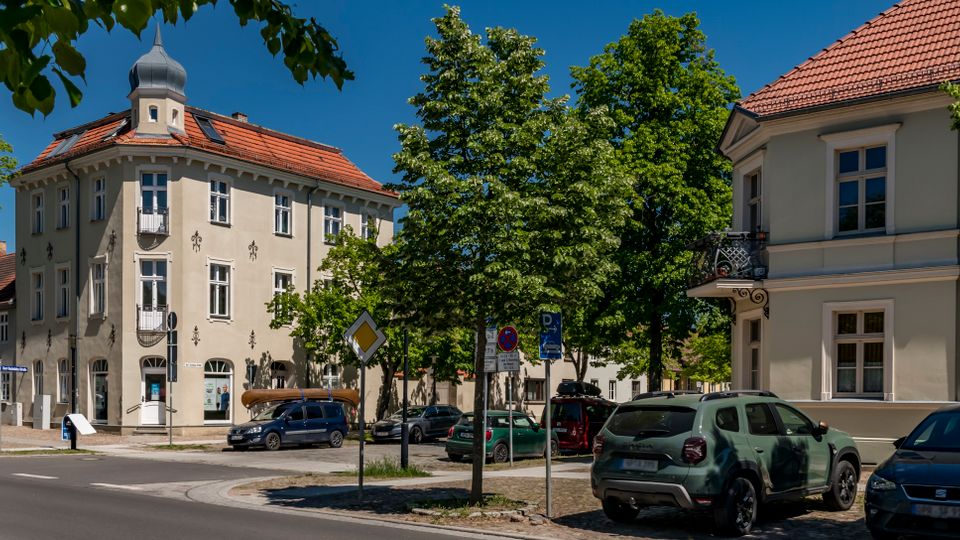 Straßenecke in einem kleinen Ortszentrum mit mehrstöckigen, hell verputzten Altbauten und roten Ziegeldächern, darunter ein Eckhaus mit kleinem Turmaufsatz. Entlang der Straße stehen parkende Autos, mehrere Verkehrsschilder und dichte Bäume, die Teile der Fassaden und den Gehweg säumen.