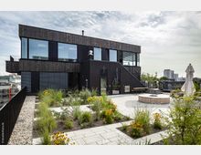 Zweigeschossiges, modernes Gebäude mit dunkler Holz- und Glasfassade, umgeben von einer Dachterrasse mit bepflanzten Beeten, Sitzbereichen und einem Sonnenschirm. Im Hintergrund sind Stadtgebäude und ein bewölkter Himmel zu sehen.