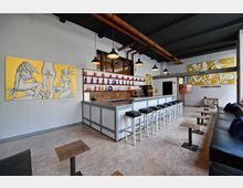 Innenraum einer modernen Bar mit grauer Theke, weißen Barhockern und einer Rückwand mit Regalen, die Flaschen und Dekor enthalten. Deckenlampen aus Metall hängen von schwarzen Balken, und an den Wänden sind großformatige, farbige Gemälde befestigt, darunter ein gelbes Bild mit abstrakten Figuren.