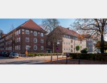 Straßenszene mit roten Backsteingebäuden im traditionellen Baustil, weißen Fensterrahmen und roten Ziegeldächern, umgeben von herbstlichem Laub und kahlen Bäumen, mit parkenden Autos und Straßenschildern im Vordergrund. Ein Gehweg und eine niedrige Hecke säumen die Straße.