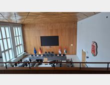 Moderner Ratssaal mit U-förmig angeordneten Tischen und Mikrofonplätzen vor einer holzverkleideten Stirnwand mit großem Bildschirm und Fahnen. Links sorgen hohe Fenster für Tageslicht, darüber spannt sich eine geometrische Holzdecke; an der rechten Wand hängt ein Wappen.