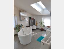 Modernes Badezimmer mit Dachschrägen, einer freistehenden weißen Badewanne, einem großen Spiegel über einem Waschtisch, einem WC, mehreren Pflanzen und einem Oberlicht, das natürliches Licht einfällt. Der Boden ist mit grauen Fliesen belegt, und ein türkisfarbener Teppich liegt vor der Badewanne.