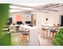 Modernes Büro mit hellen Wänden und offenen Arbeitsbereichen, ausgestattet mit Schreibtischen, Computerbildschirmen und hohen Stehtischen mit Barhockern. Sichtbare Lüftungsrohre an der Decke und Dekorationen wie ein großformatiges Gemälde an der Wand ergänzen den Raum.