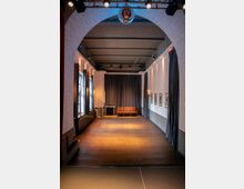 Ein schmaler Raum mit Holzboden und hohen Decken, dekoriert mit Vorhängen an den Wänden. Im Hintergrund steht ein braunes Ledersofa vor einem geschlossenen Vorhang, und an den Seiten hängen gerahmte Bilder.