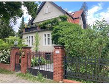 Zweistöckiges Wohnhaus im traditionellen Stil mit hell verputzter Fassade, roten Ziegeldächern und einem Balkon, umgeben von dichtem Grün und einem gepflegten Garten. Die Einfahrt ist durch ein schmiedeeisernes Tor und eine Mauer aus roten Ziegeln mit dekorativen Elementen eingefasst.