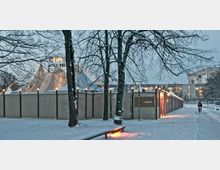 Schneebedeckter Platz mit einem großen Zirkuszelt, das hinter einer Holzwand sichtbar ist, verziert mit Lichterketten. Im Vordergrund stehen schneebedeckte Bäume, während im Hintergrund ein modernes Gebäude erkennbar ist.