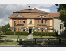 Fassade eines zweistöckigen Restaurants mit orange-rotem Anstrich und einem Dach aus roten Ziegeln, beschriftet mit "Zum alten Schloß". Vor dem Gebäude befinden sich Grünflächen, große Werbetafeln und mehrere beschattete Sitzbereiche mit grünen Sonnenschirmen.