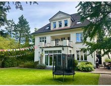 Großes zweistöckiges Einfamilienhaus mit weißer Fassade und Satteldach, umgeben von einem gepflegten Garten mit grünem Rasen, Sträuchern und Bäumen. Im Vordergrund ist ein Trampolin zu sehen, und bunte Wimpelketten schmücken den Garten und die Terrasse.