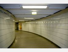 Unterirdischer Fußgängertunnel mit gekachelten weißen Wänden, beleuchtet von rechteckigen Deckenleuchten. Im Hintergrund ist eine orangefarbene Werbetafel erkennbar.