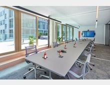 Moderner Konferenzraum mit einem langen Tisch in der Mitte, umgeben von grauen Bürostühlen. Große Fenster auf der linken Seite lassen viel Tageslicht herein, während die rechte Wand einen großen Bildschirm zeigt; Pflanzen und Schreibutensilien dekorieren den Raum.