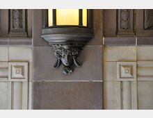 Detailaufnahme einer Wand mit dekorativen Elementen aus Stein, darunter ein Relief einer Frauenmaske mit geflochtenem Haar und Blumenmotiv, das die Basis einer antiken Lampe mit warmem Licht bildet. Die Wand zeigt quadratische Fliesen mit kassettenförmigen Verzierungen.