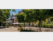 Ein gepflasterter Platz mit Bäumen, die Schatten spenden, umgeben von gelben Gebäuden und einem modernen Anbau mit Glasfassade. Im Vordergrund stehen Tische und Sonnenschirme eines Cafés oder Restaurants, im Hintergrund ist das Gebäude eines Theaters oder Kulturzentrums zu sehen.