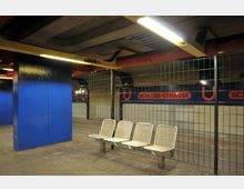 U-Bahn-Station mit einem blauen Pfeiler, einem orangefarbenen Lüftungsrohr unter der Decke und vier Metallbänken vor einem grünen Gitterzaun. Im Hintergrund ist ein Schild mit der Aufschrift „Schlossstraße“ und einem U-Bahn-Symbol an der Wand zu sehen.