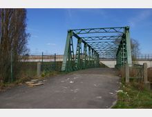 Grüne Stahlbrücke mit Fachwerkstruktur über einer Straße, umgeben von einem Maschendrahtzaun und kahler Vegetation; Asphaltweg führt zur Brücke, im Hintergrund blauer Himmel und einige Bäume.