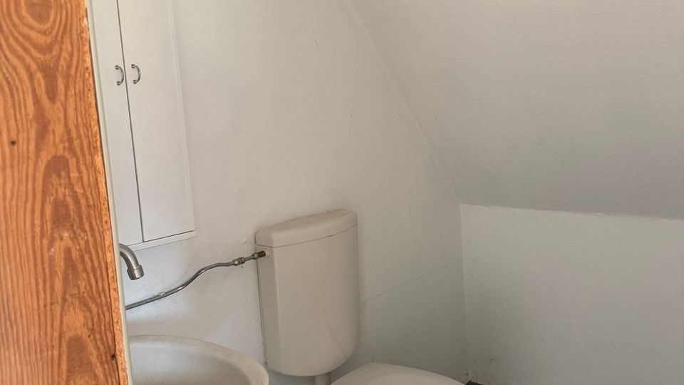Kleines WC unter einer Dachschräge mit weiß gestrichenen Wänden, Toilette mit Spülkasten und einem kompakten Waschbecken neben einem schmalen Wandschrank. Dunkler Boden aus unregelmäßigen Fliesenfragmenten und Holztürrahmen im Vordergrund.