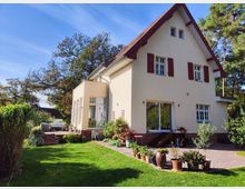 Ein zweistöckiges Einfamilienhaus mit Satteldach, beigefarbener Fassade und roten Fensterläden, umgeben von einem gepflegten Garten. Im Vordergrund ist eine Terrasse mit Pflanzentöpfen und Gartenmöbeln zu sehen, daneben eine große Rasenfläche und Bäume im Hintergrund.